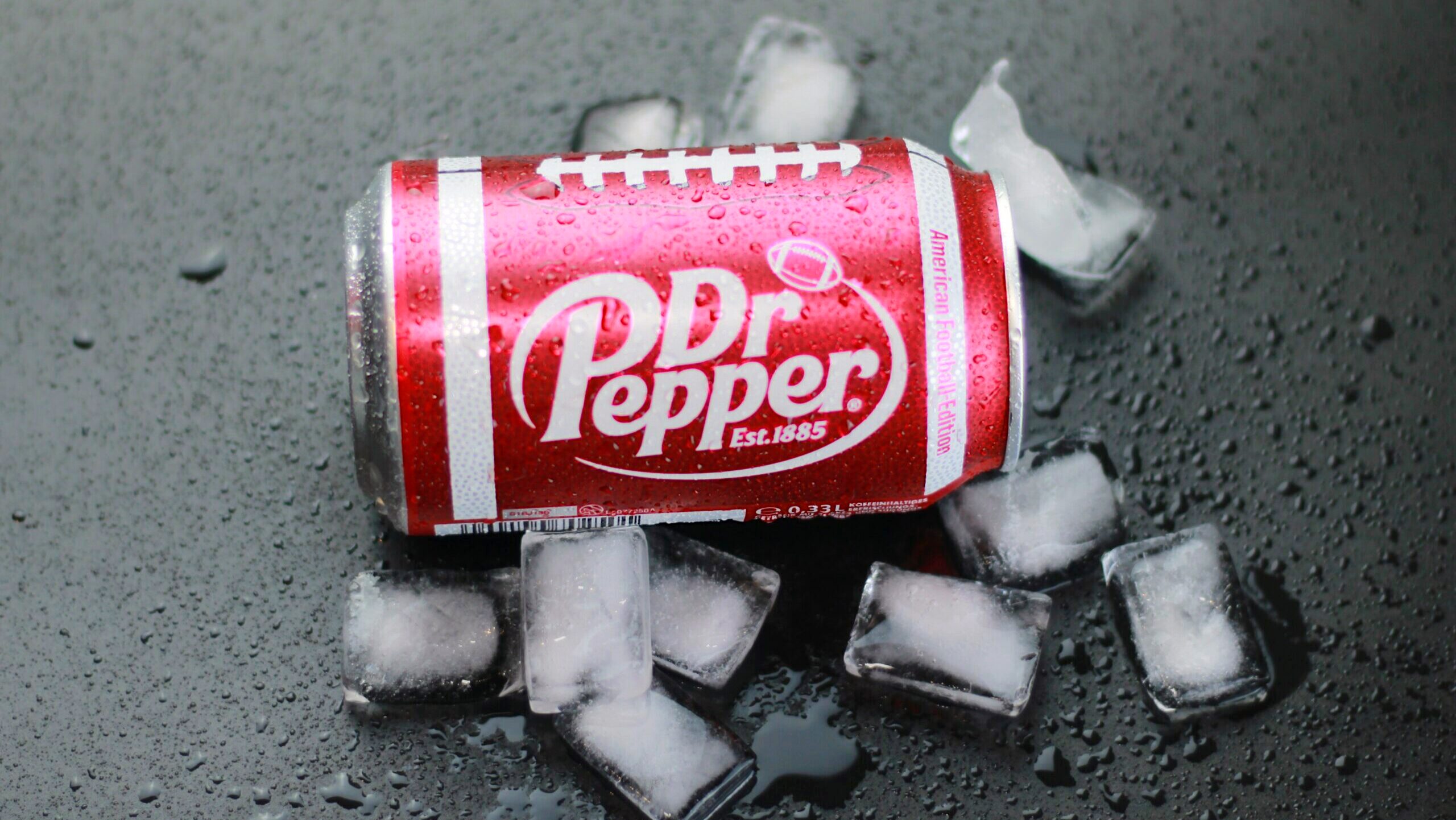 A chilled can of Dr. Pepper soda is shown laying among ice cubes on a flat surface.