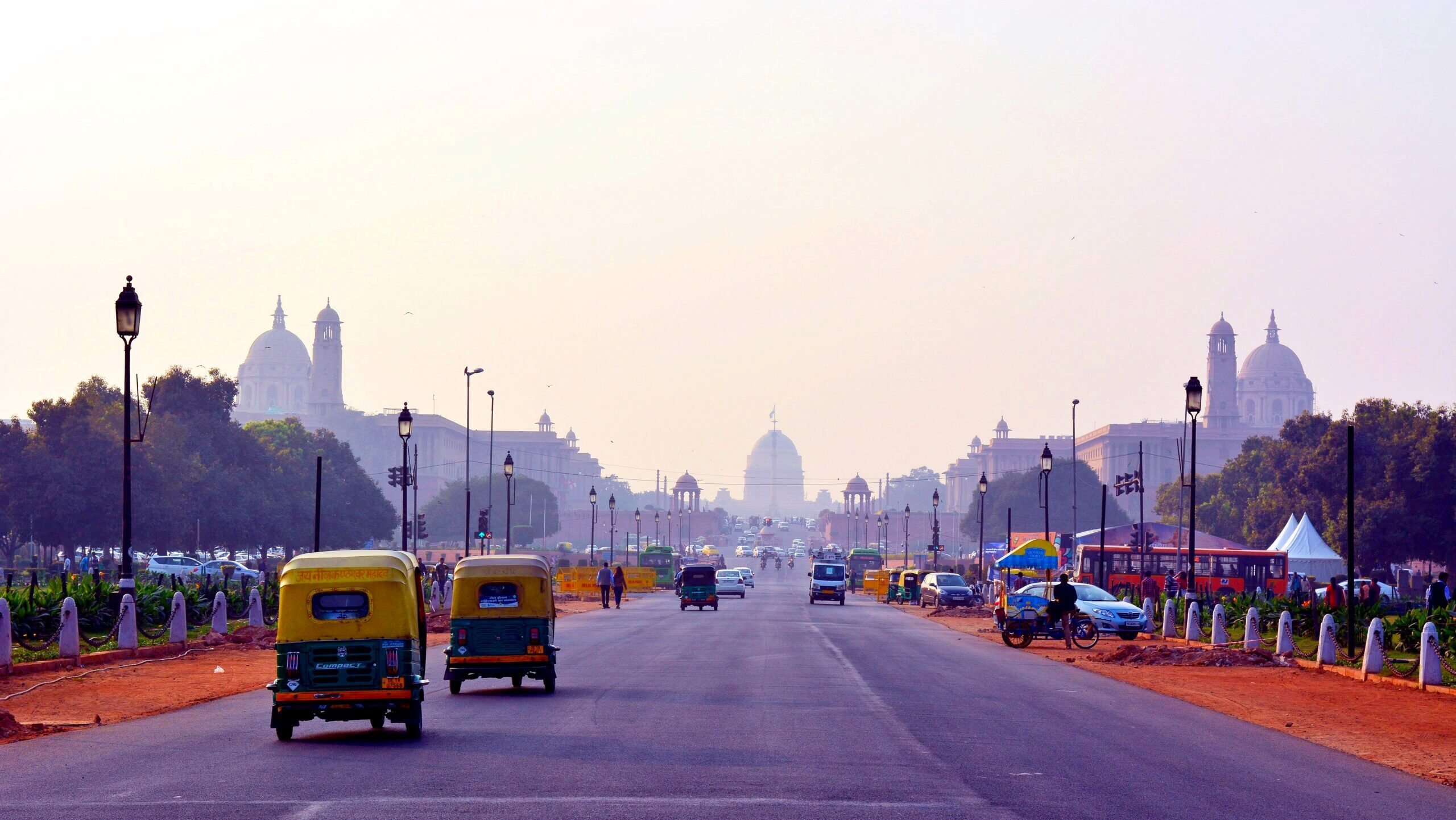 Taxis drive down a boulevard in New Dehli leading up to a complex of government buildings.